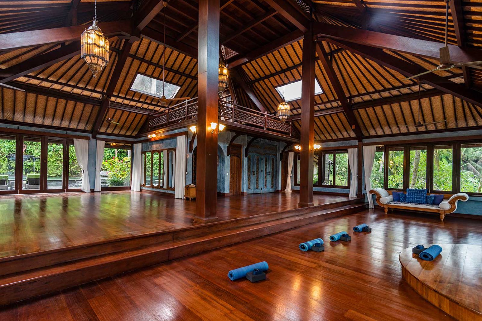 Inside the yoga shala, a vaulted timber ceiling and polished hardwood floor with mats laid out and windows open to the trees